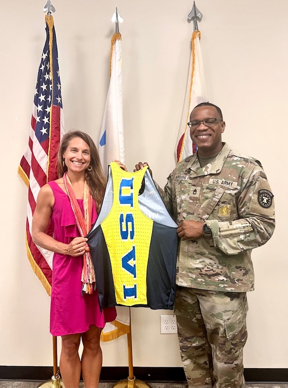 Man standing on the right in an Army uniform and woman standing on the left in a pink dress hold a USVI jersey.