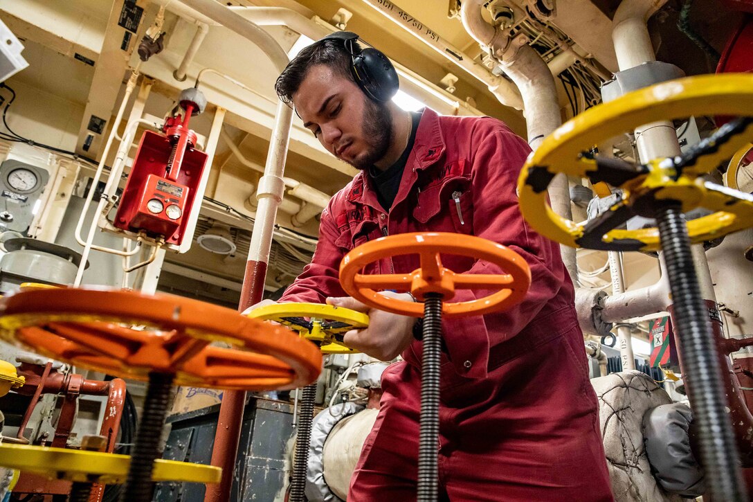 A sailor wearing ear protection turns a control wheel inside an equipment room on a ship.