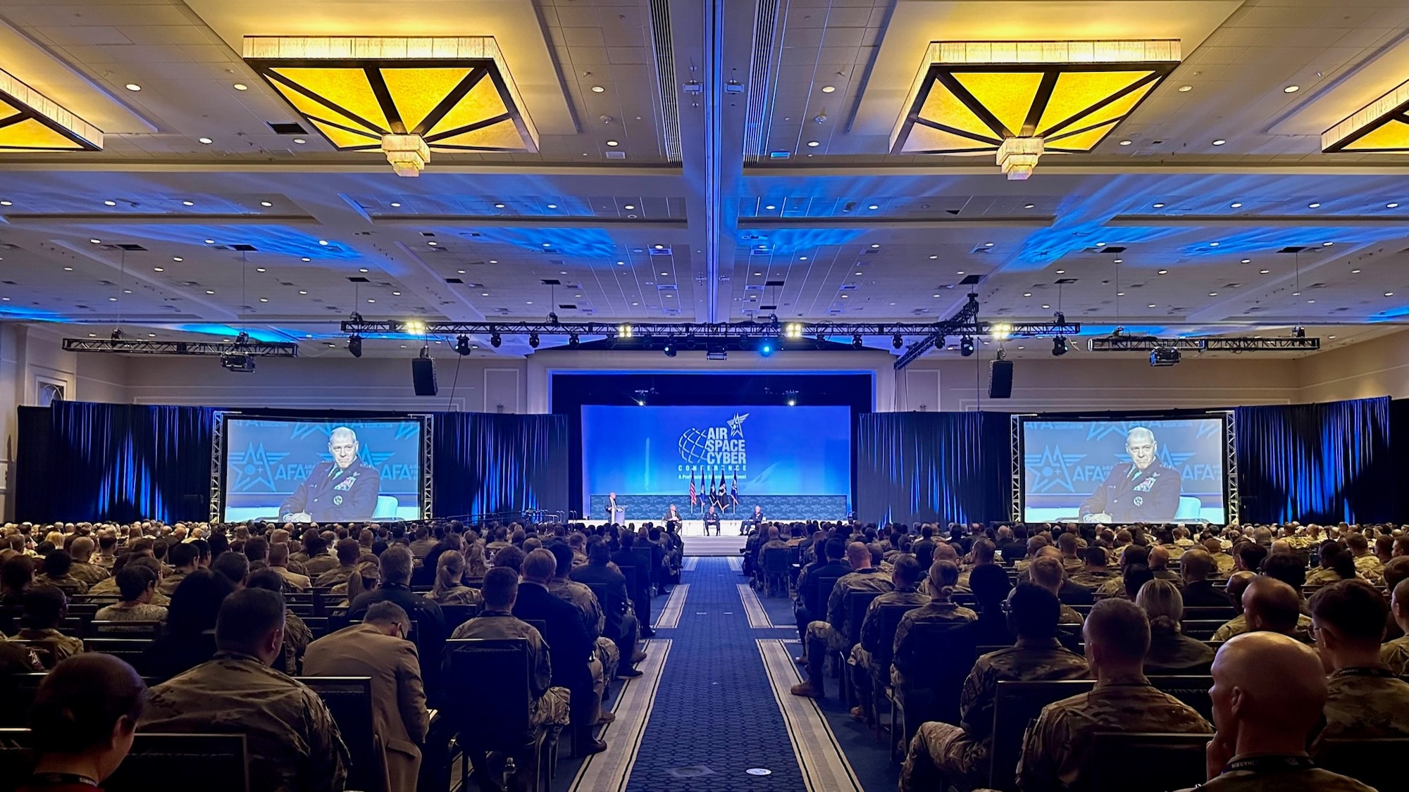 The photo is the perspective from the back of an auditorium which is filled with many people. The stage has people on it. There are two large projector screens on either side of the stage which show the zoomed in image of AFGSC commander, General Thomas Bussiere, as he sits on the center stage with the other people. This is during a conference.