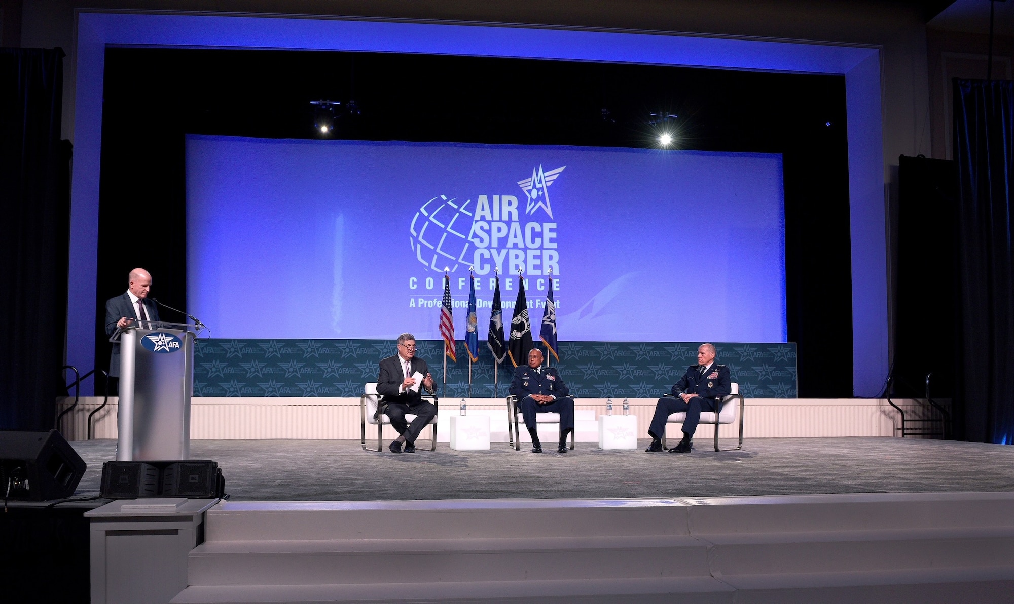 Three people sit in chairs next to a someone speaking at a podium. They are all on a stage during a conference.