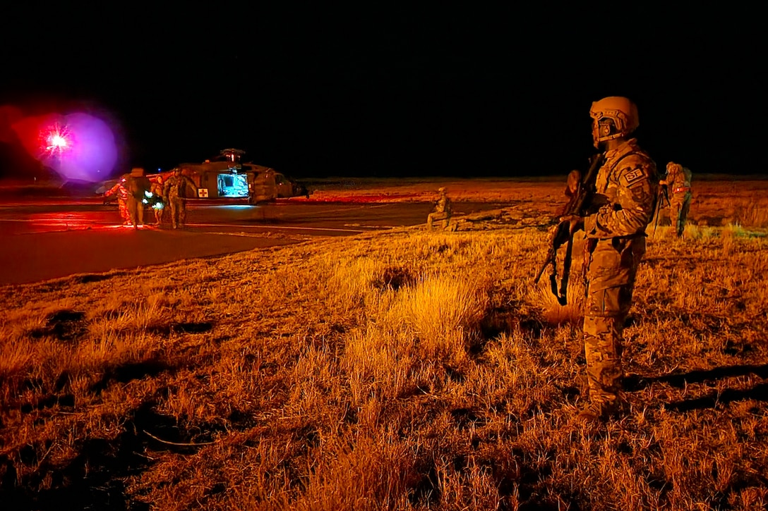 Airmen carrying weapons watch as fellow airmen run toward a helicopter carrying a simulated patient in the dark illuminated by a spotlight.