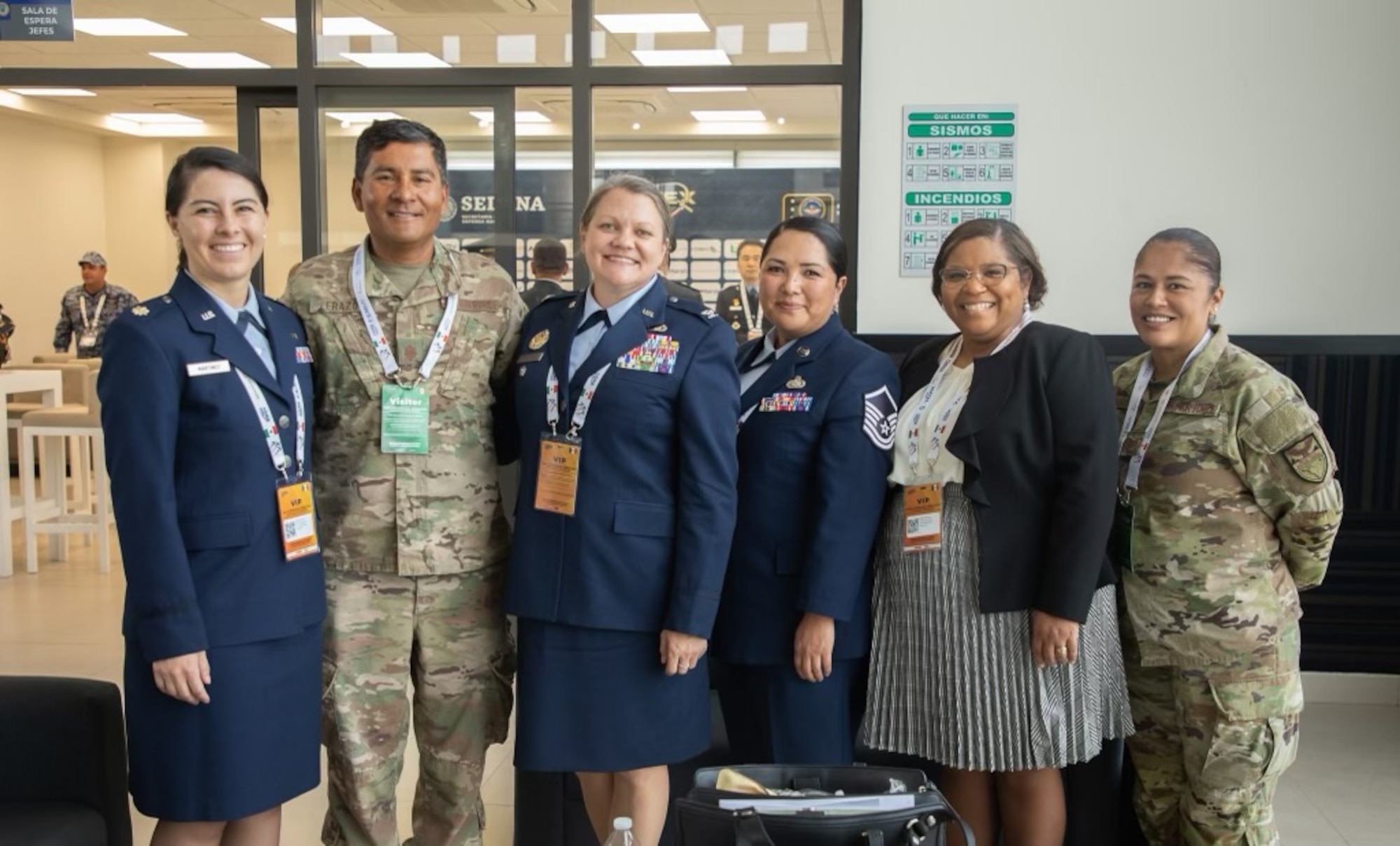 A group of people in a mixture of U.S. Air Force service dress, operational camouflage and civilian attire stand in a line for a group photo.