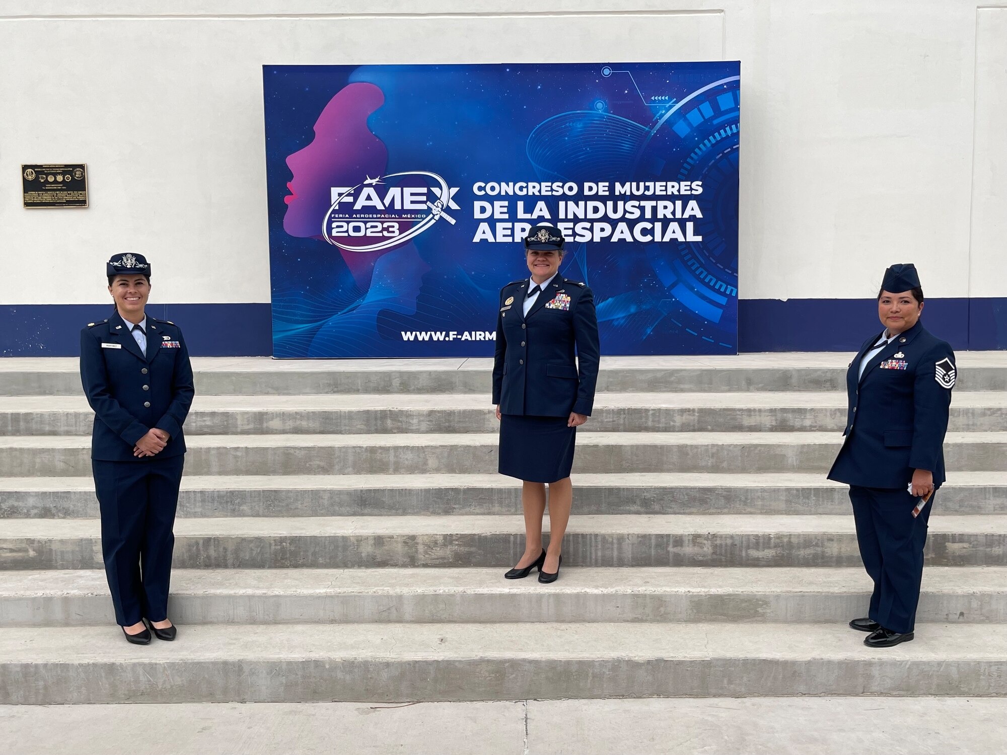 Three women in U.S. Air Force service dress stand on a staircase with a sign in the background that reads "Congreso de Mujeres de la Industria Aeroespacial FAMEX 2023"