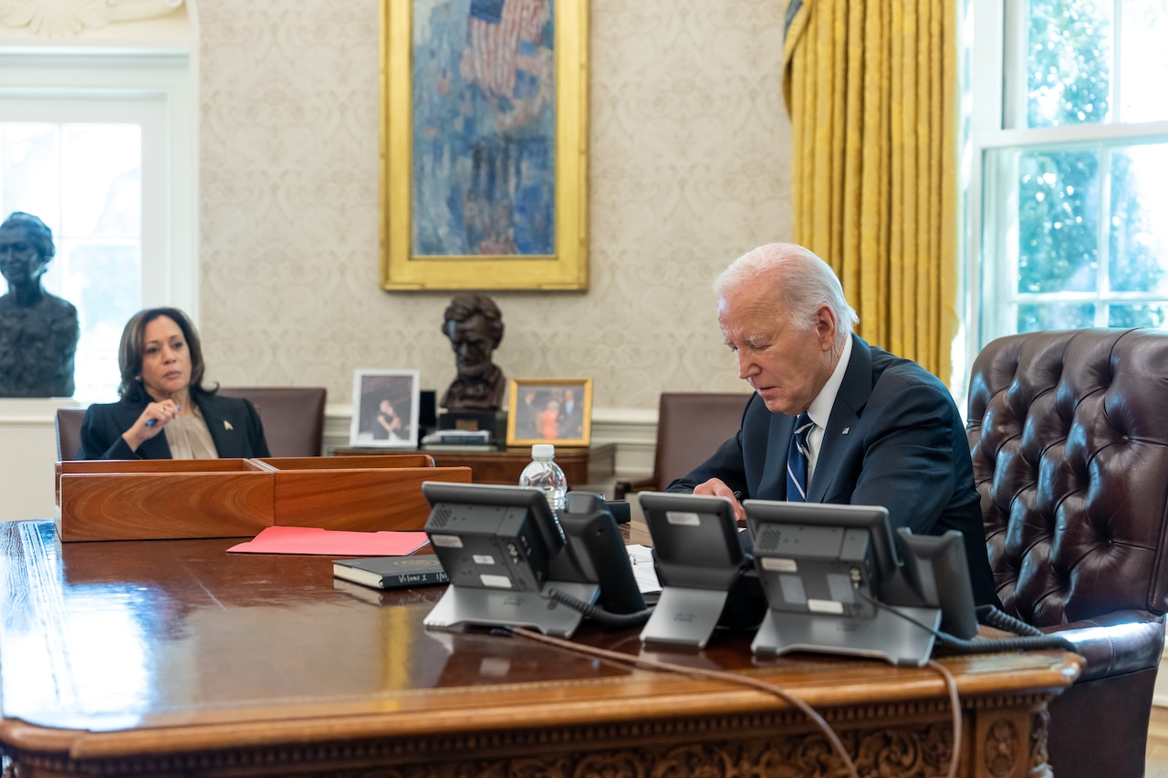 President Joe Biden and Vice President Kamala Harris across from each other at a desk in front of three telephones.