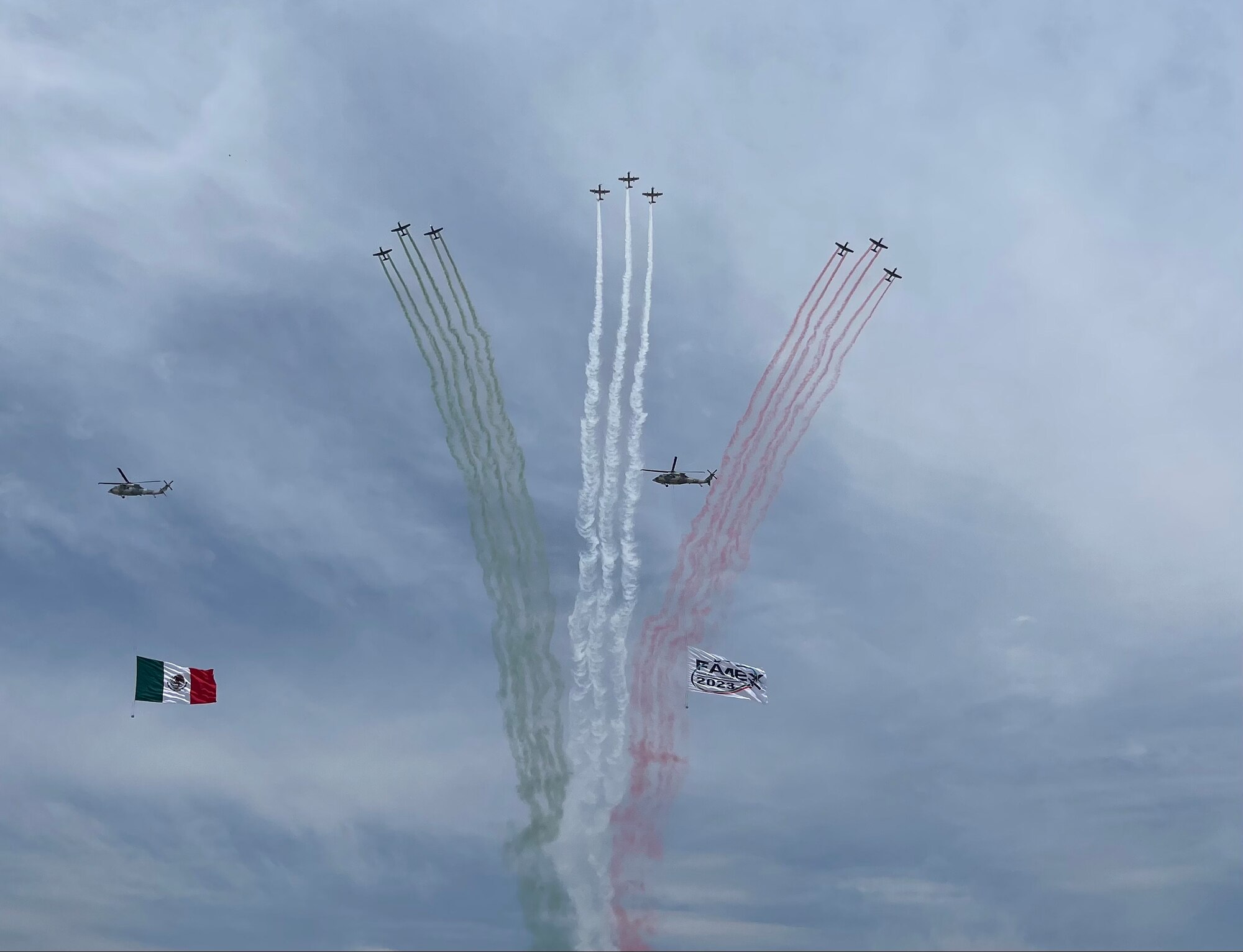 9 aircraft in formation create smoke streams -- 3 red, 3 white, 3 green -- as 2 helicopters fly through the scene, another aircraft not visible flying the Mexico flag and a FAMEX sign through the sky.