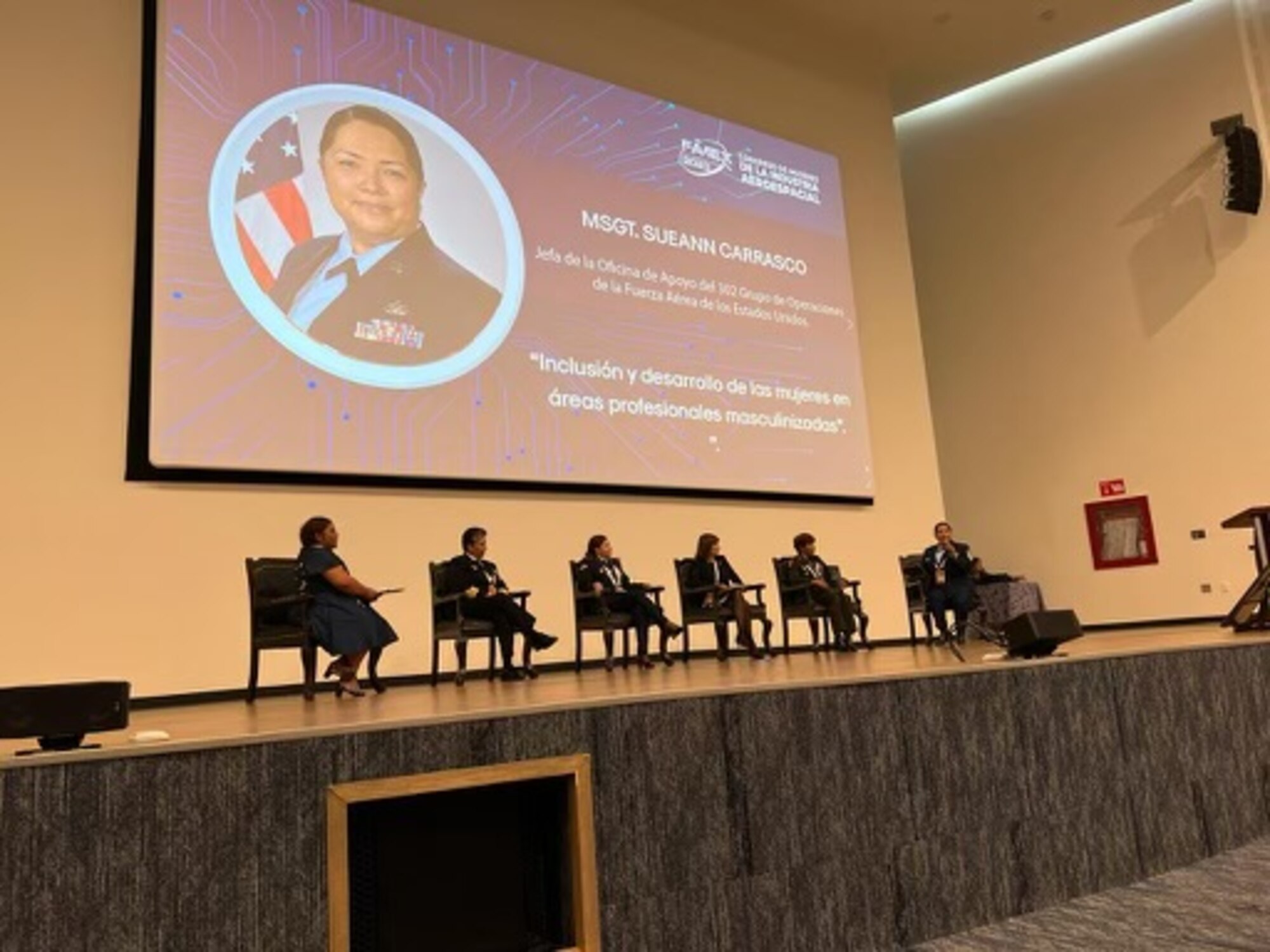 A row of panelists speaks on a stage with a screen behind them displaying a portrait of one of the speakers, Master Sgt. Sueann Carrasco.