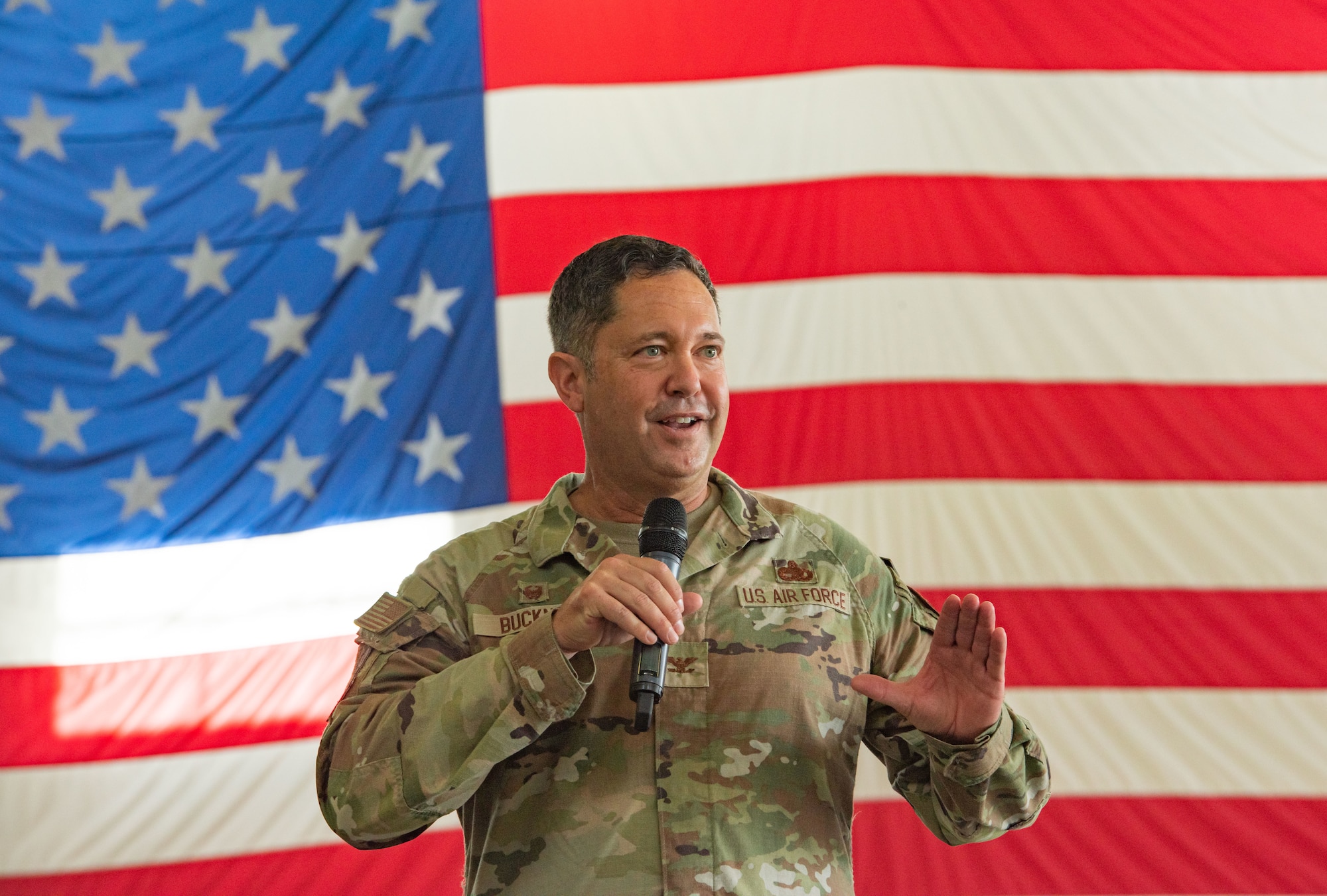 Col. Bobby Buckner holds a microphone and gives a speech in front of a U.S. flag.