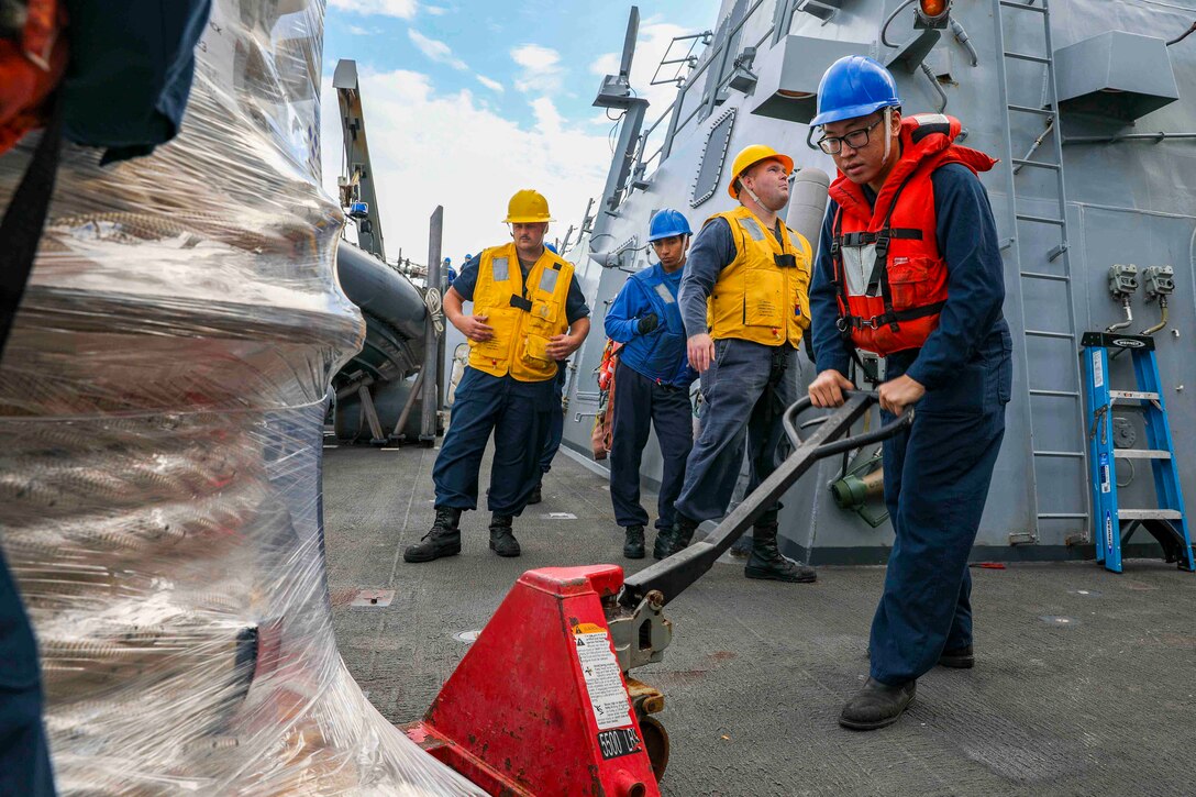 A sailor uses a pallet jack to move a load of supplies on the deck of a ship as other sailors stand behind.