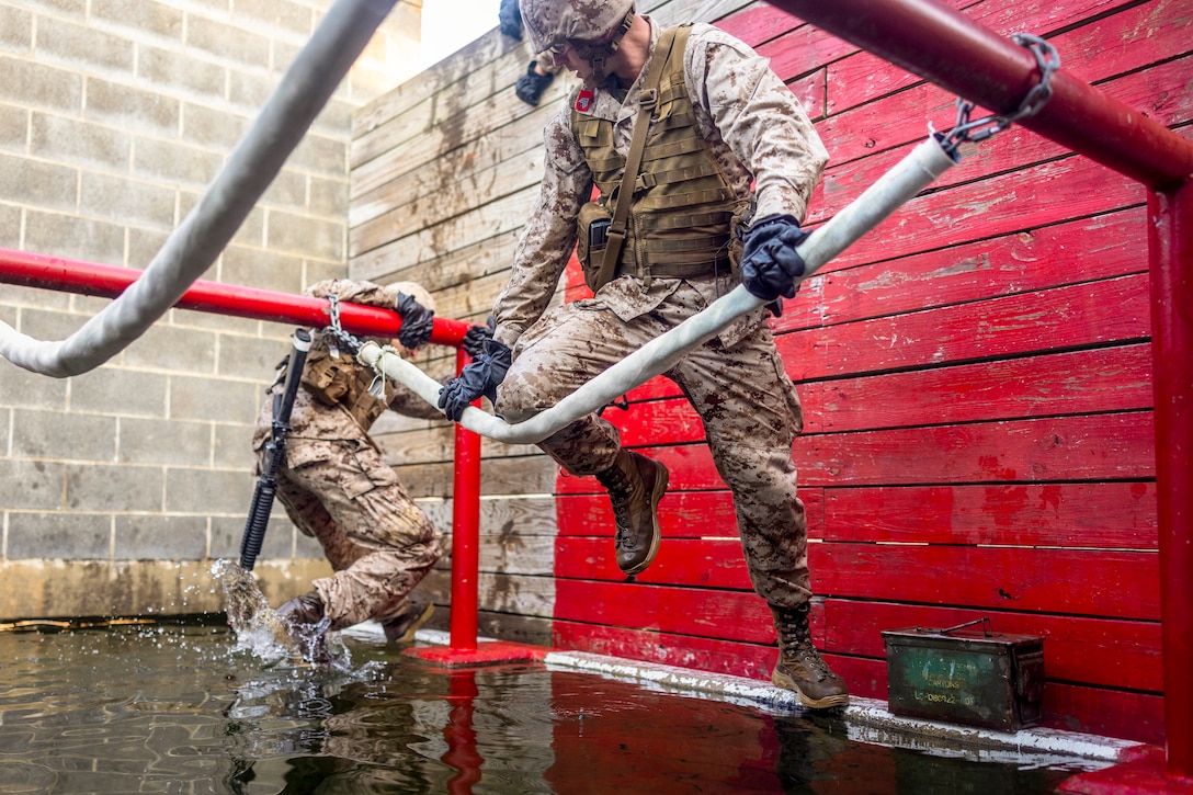 A Marine Corps officer candidate climbs on a chain attached to two red poles as a fellow candidate jumps out of a puddle of water to begin the obstacle course.