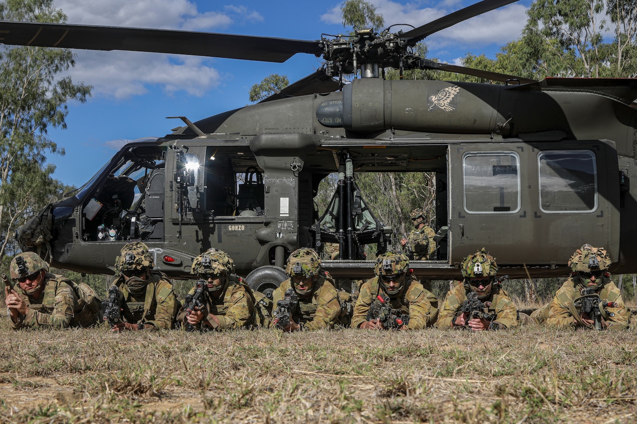 Soldiers lie on their stomachs with weapons pointed in front of a helicopter.