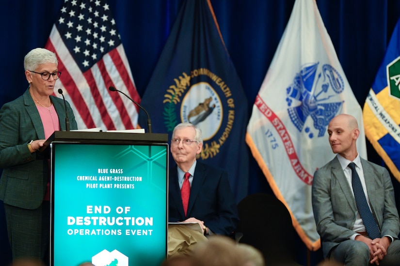 A person wearing glasses stands at a lectern and speaks into a microphone as two seated people look on.