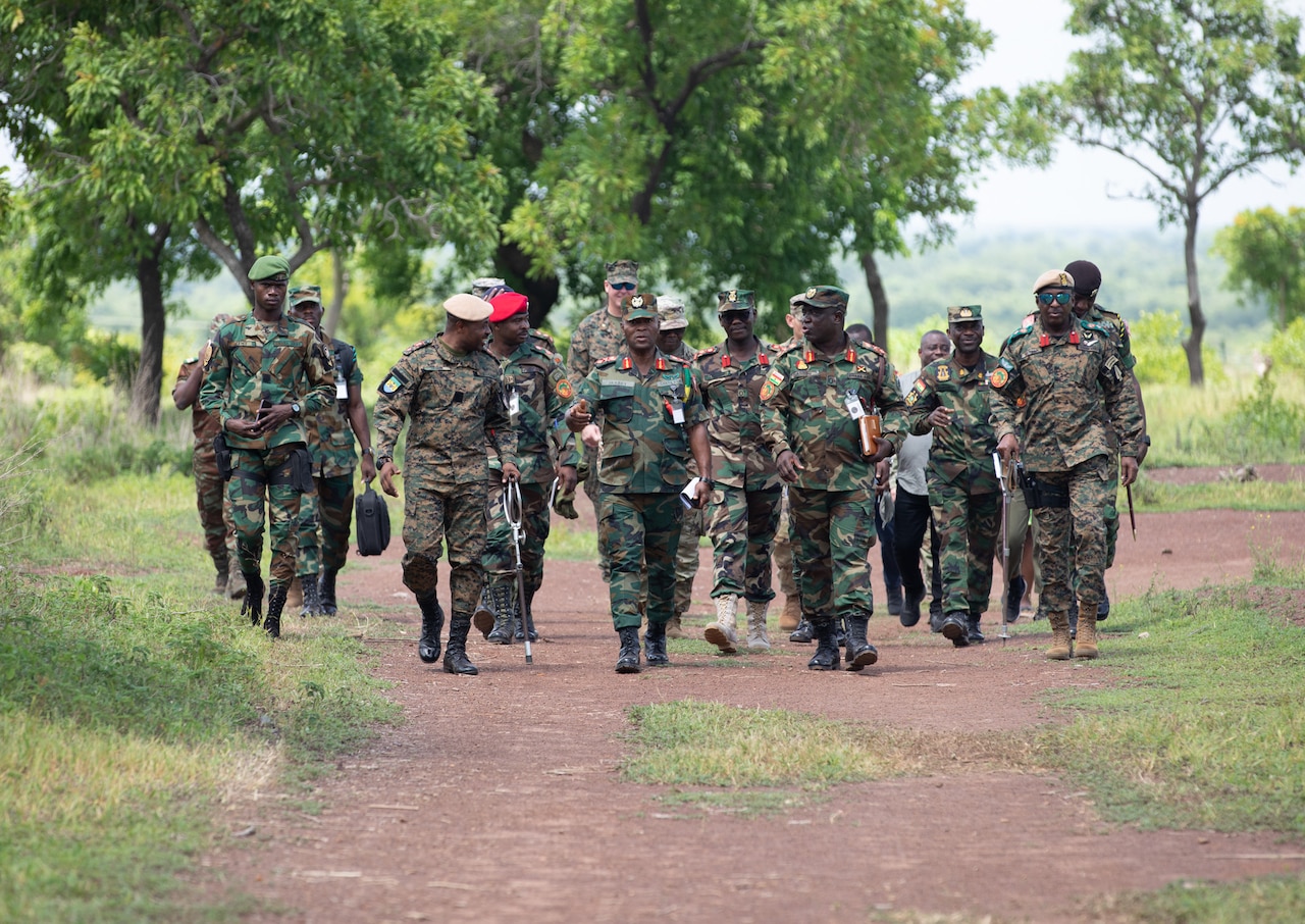 Ghana military officers walk in formation on a dirt road surround by a wooded area.