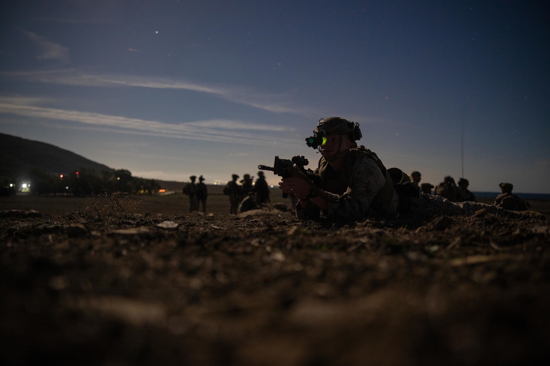 A Marine lies on the ground at twilight holding a weapon with several other Marines in the background.