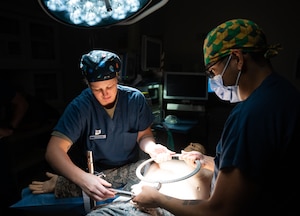 A female Airman in scrubs holds metal ring with a male Airman in scrubs over a training dummy