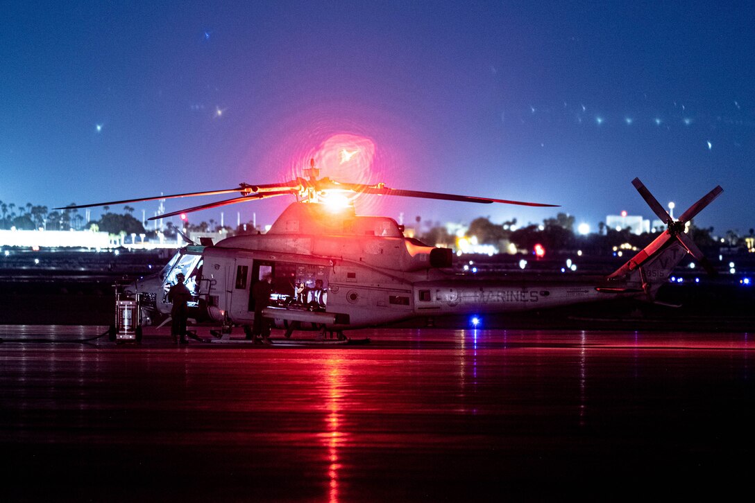 A military helicopter is parked on the tarmac at night. The photo is illuminated by a red light atop the helicopter.
