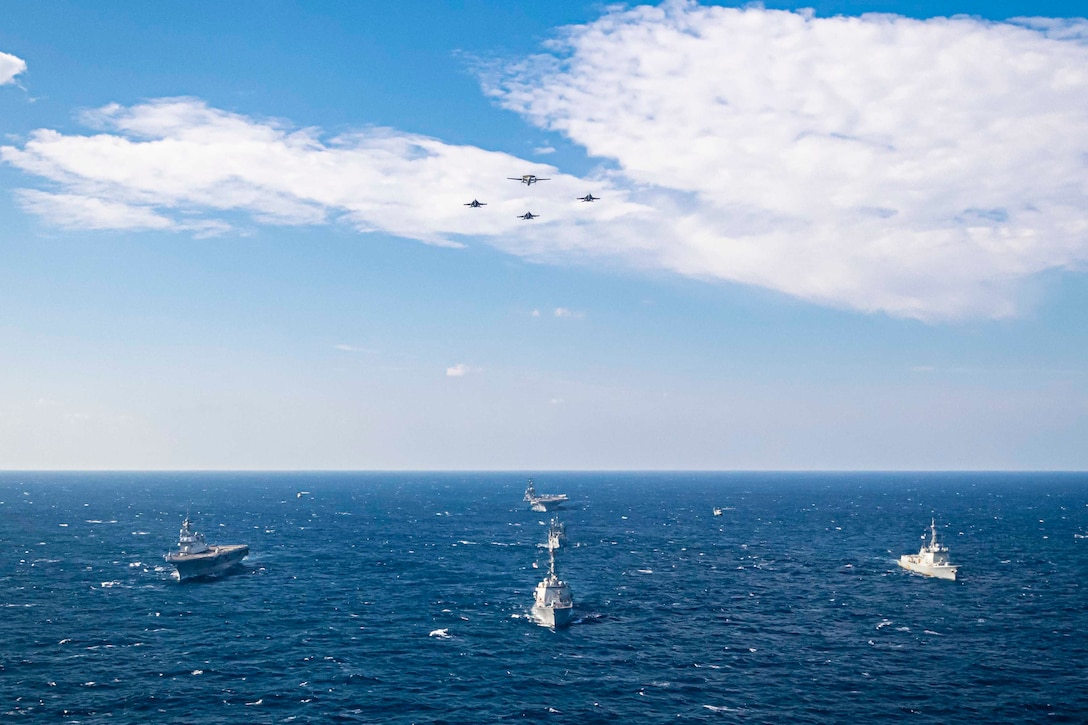 Five ships sail in formation through a body of water as four aircraft fly above.