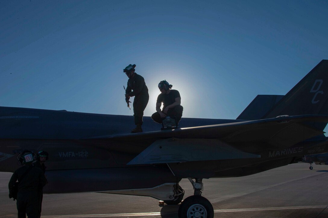 Two Marines shown in silhouette stand on the wing of a parked aircraft while talking to two others standing below.