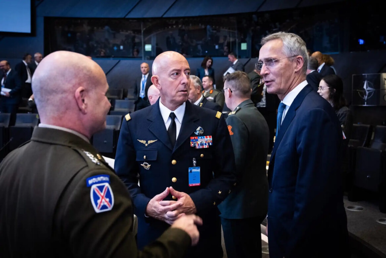 Two men in military uniform, speak with a man in business attire.