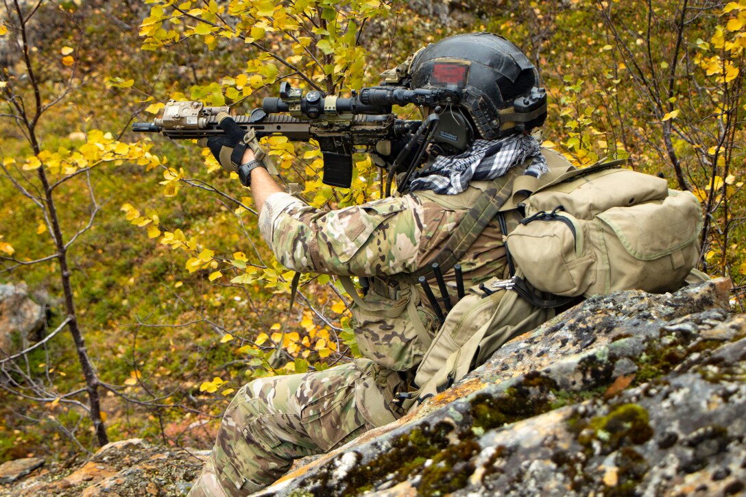 A soldier in camouflage aims a weapon while leaning on a rock covered in moss in a wooded area.