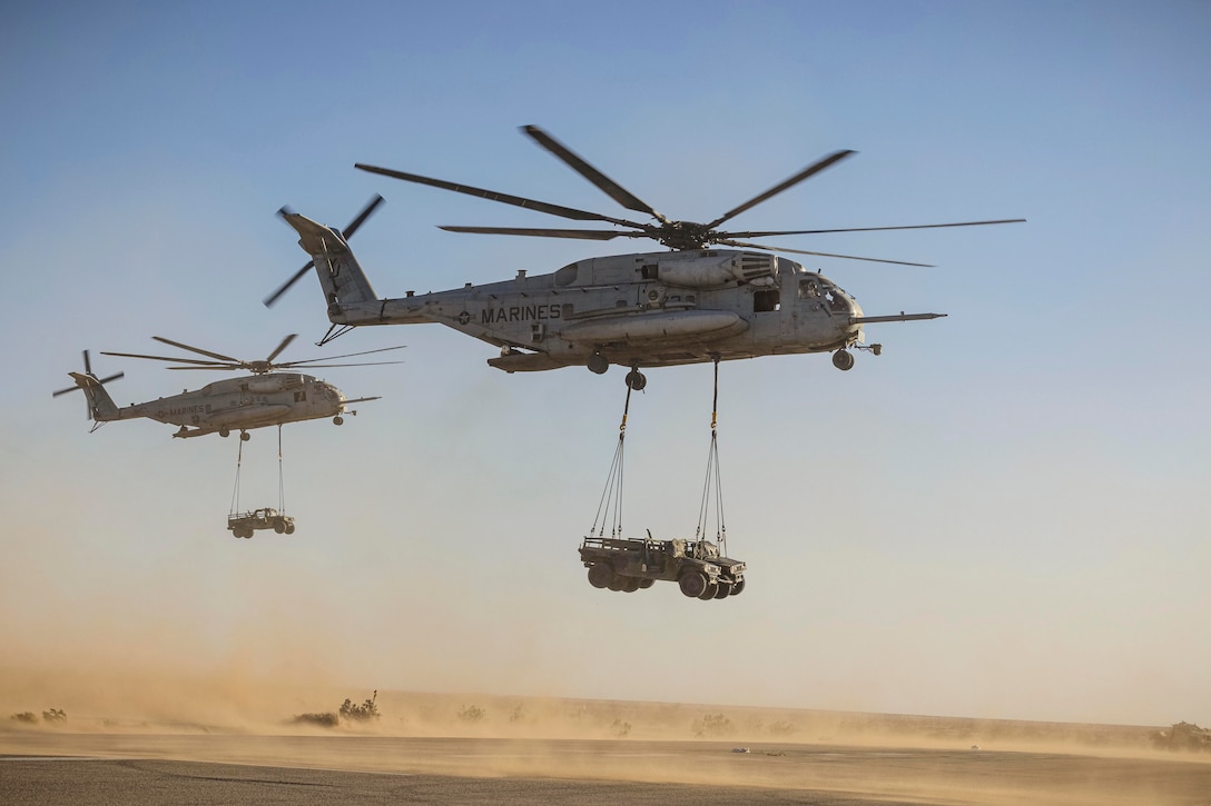 Service members ride in Humvees in a desert-like area as a Humvees hoisted to two helicopters fly above.