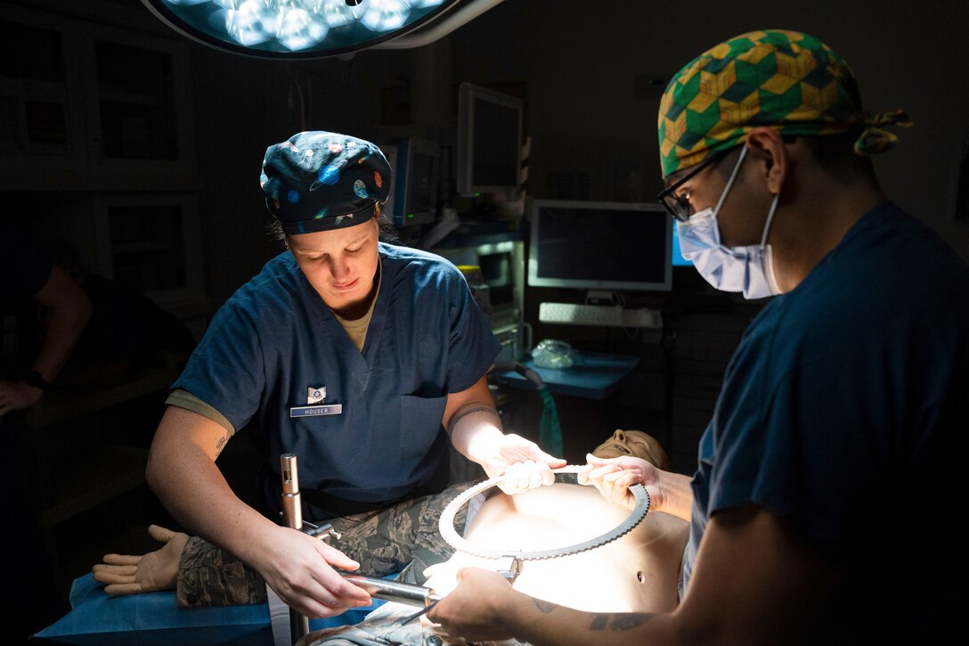 Airmen disassemble a table-mounted retractor from over a dummy laying on an operating table illuminated by a spotlight.