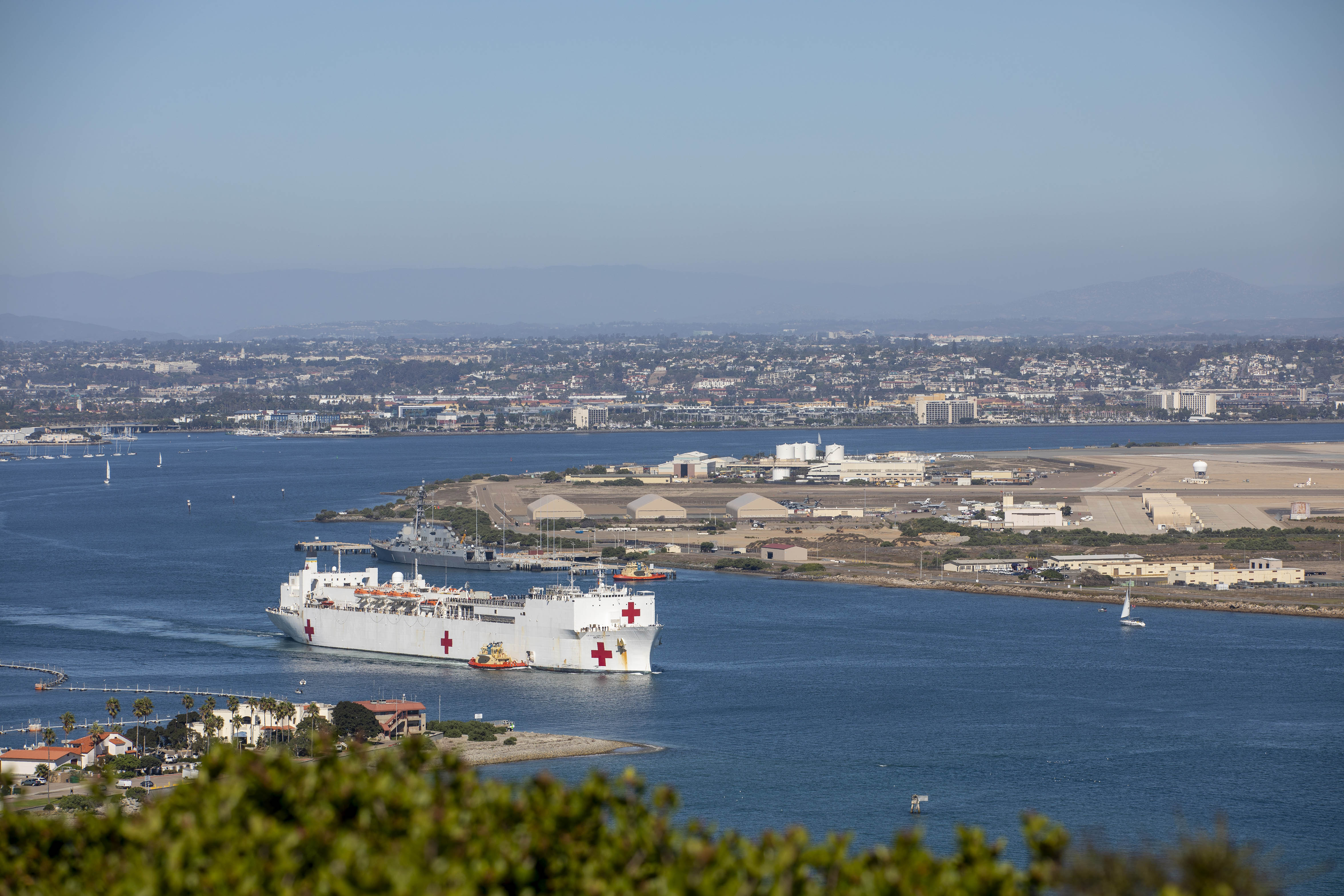 A Navy ship with red crosses along the side and rear sails from a port.