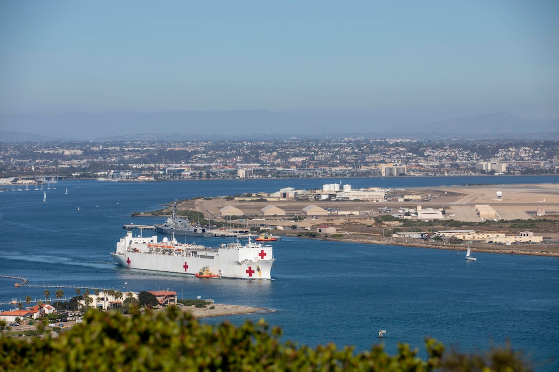 A Navy ship with red crosses along the side and rear sails from a port.