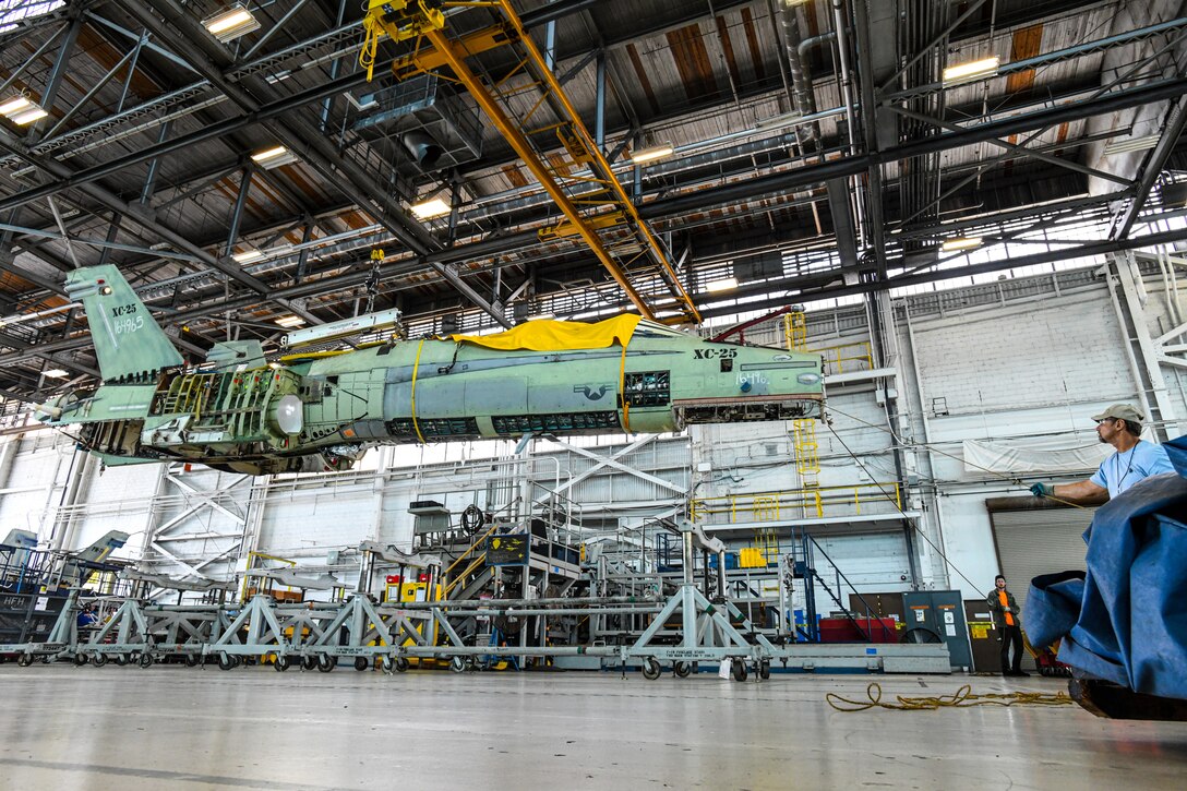 A Fleet Readiness Center Southeast (FRCSE) weight and handling crewmember helps to stabilize an F/A-18 Hornet as it is repositioned using a crane as part of a Center Barrel Replacement (CBR) maintenance evolution. CBRs were conducted to extend the service life of F-18A-D aircraft – saving the U.S. government millions in replacement costs. (U.S. Navy Photo by Toiete Jackson/Released)