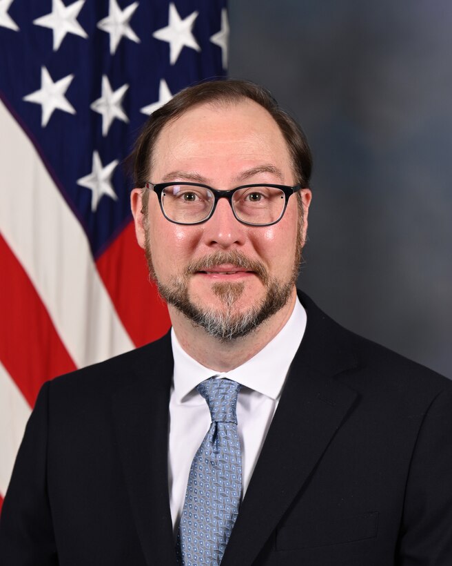 A man in a suit poses for a photo in front of the American flag.