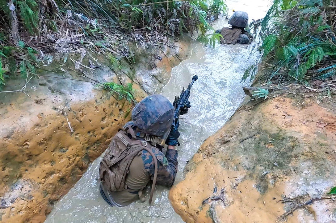 Marines move through a water-filled trench while holding weapons.