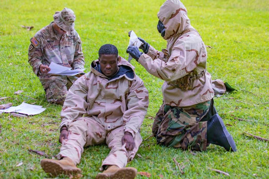 A soldier sits between two other soldiers kneeling; one is looking through papers and the other prepares bandages.