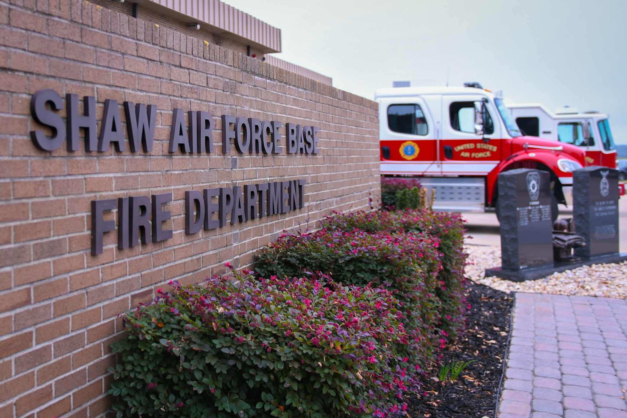 The photo is taken at the entrance of a fire department sign that states, "Shaw Air Force Base Fire Department." There are two fire trucks parked in the back ground.