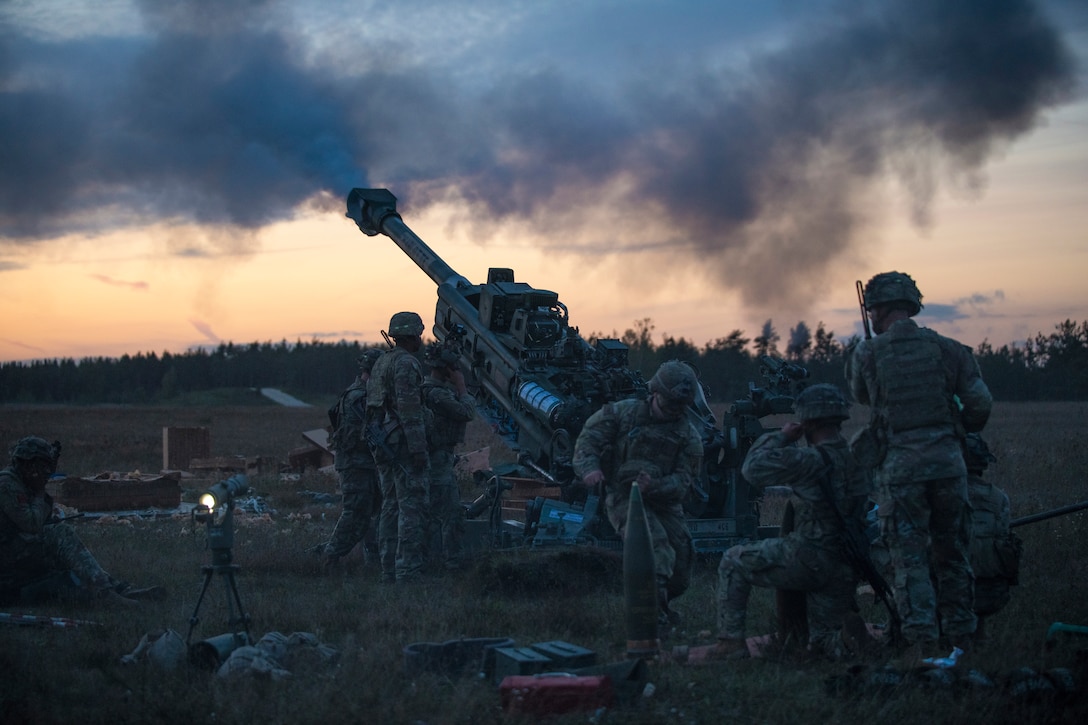 Smoke fills the air as soldiers stand or kneel near a recently fired weapon in a field covered with training supplies.