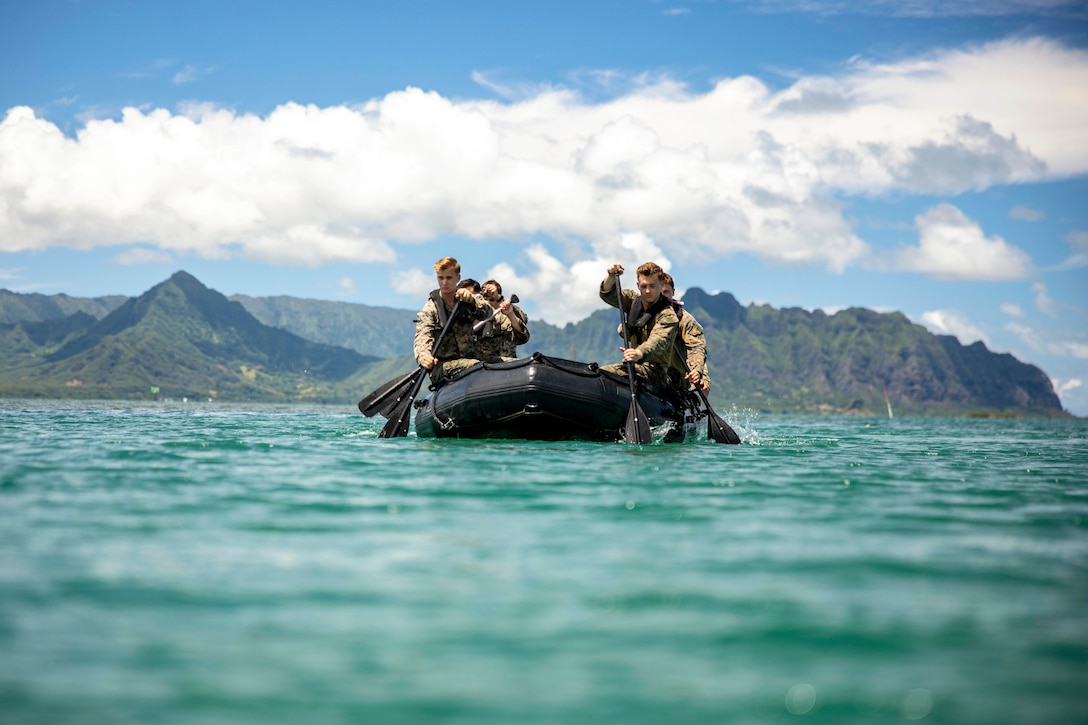 Marines paddle in a small rubber boat with mountains and partly cloudy skies in the background.