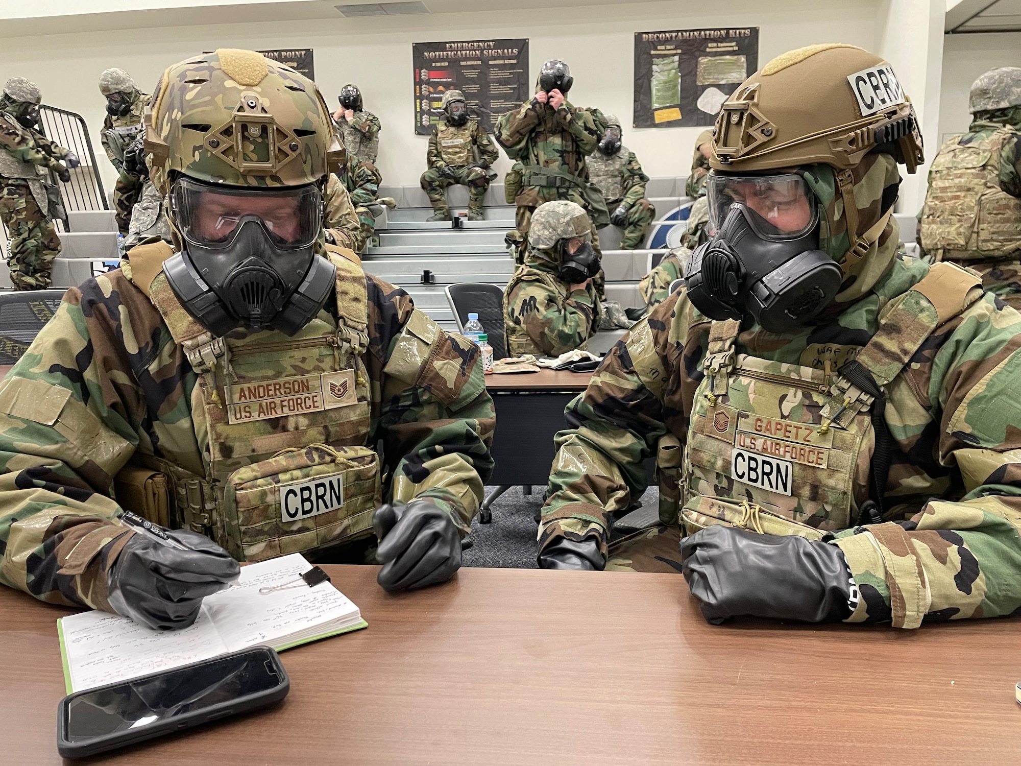 two soldiers clad in full protective gear and gas masks write notes at a table