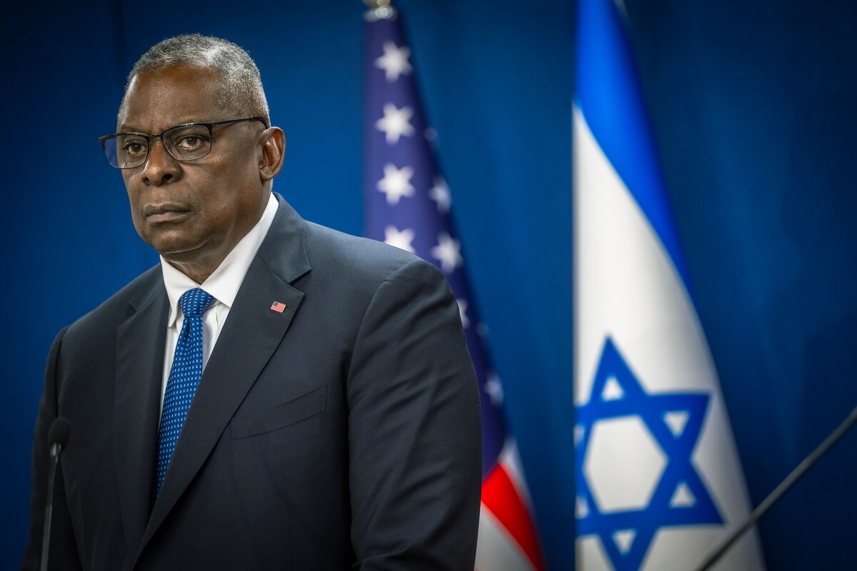 A man in a suit stands at a lectern with U.S. and Israeli flags in the background.