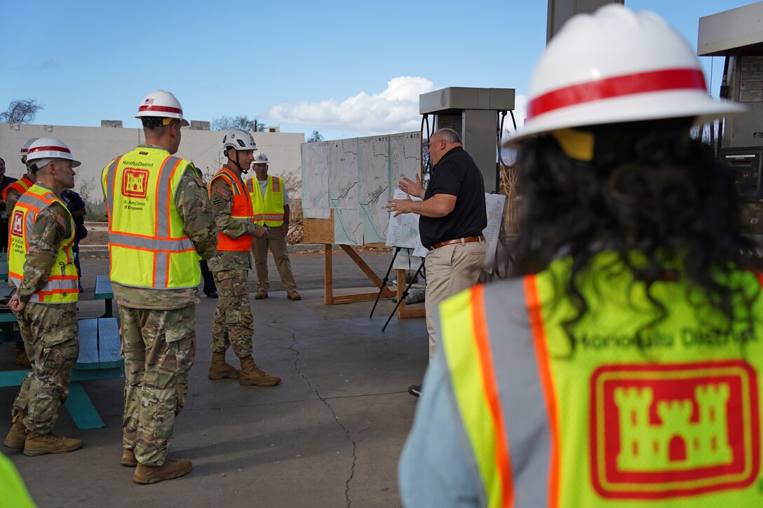 Several people wearing safety vests and hardhats receive a briefing from a man in an outdoor setting.