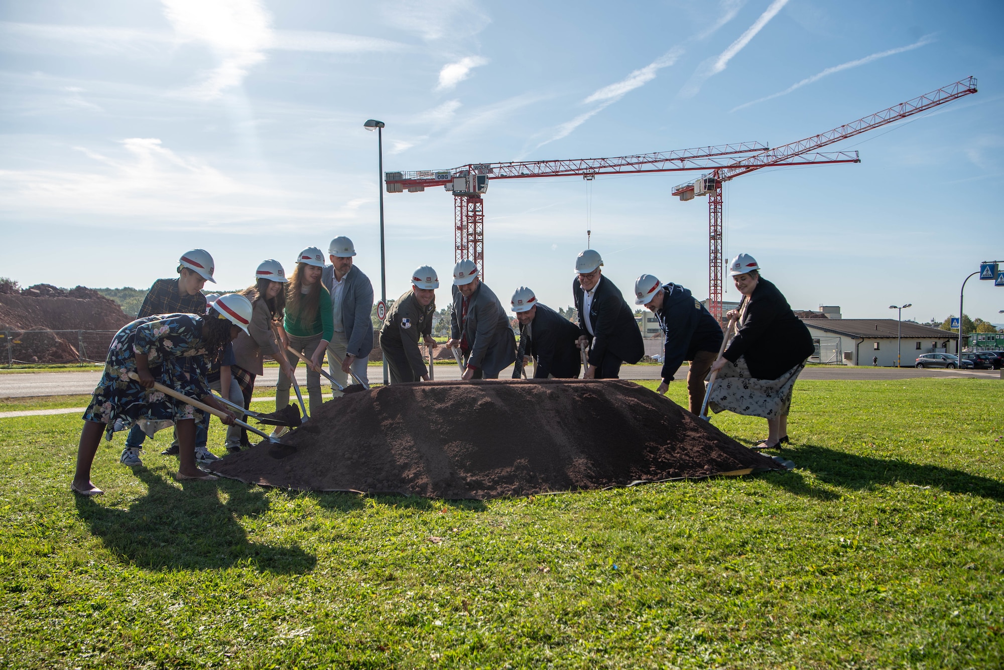 Military and civilian leaders and students wearing hard hats shovel a pile of dirt in front of cranes and construction.