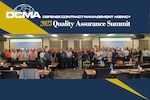 Group photo of several people standing in front of projectors. Text on graphic reads Defense Contract Management Agency 2023 Quality Assurance Summit.