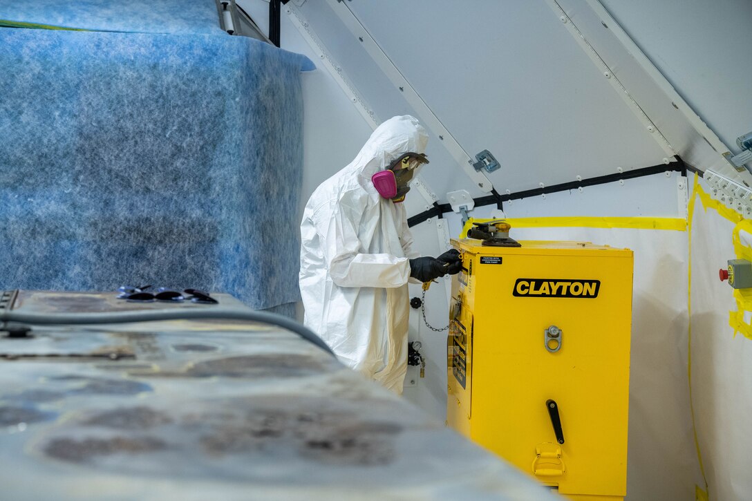 An airman in a protective suit handles tools in a mobile paint booth.