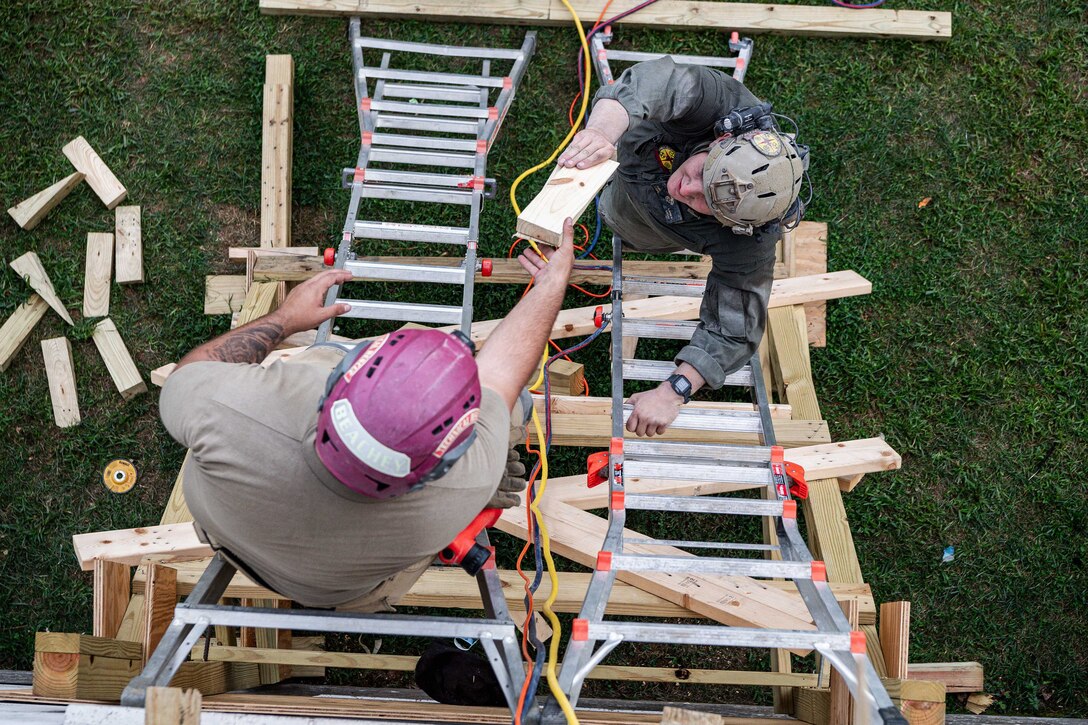 A service member on a ladder passes a piece of wood to another on a ladder.