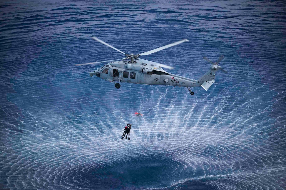 Sailors are hoisted by an airborne aircraft over a body of water.