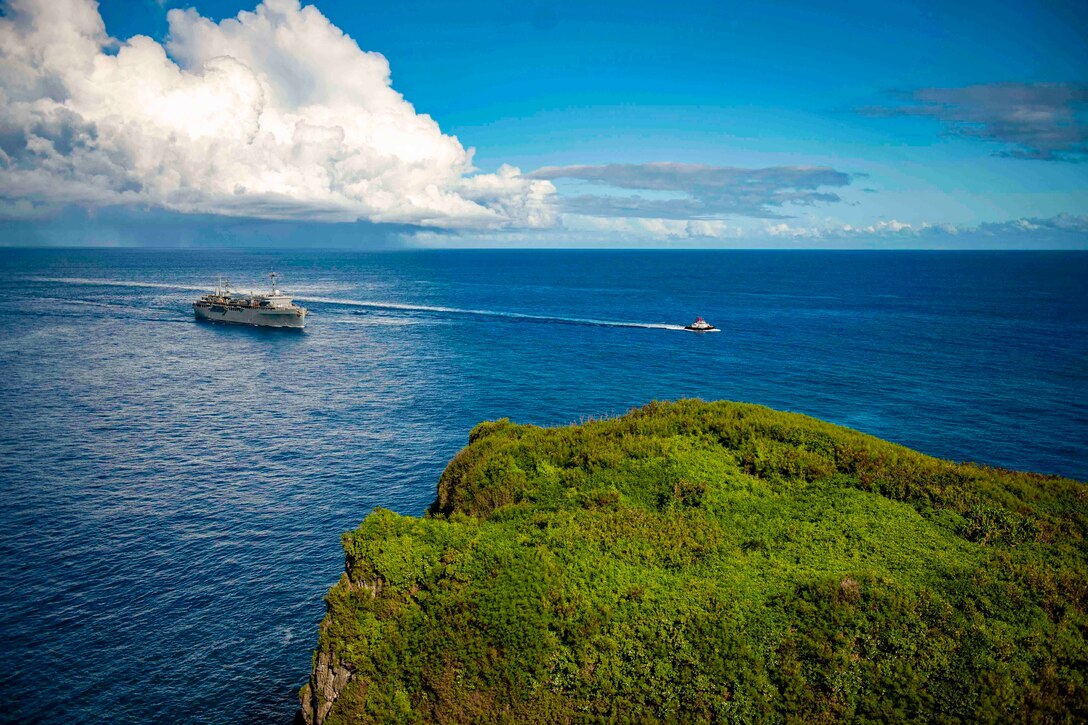 A ship and a boat travel through a body of water next to a coastline.