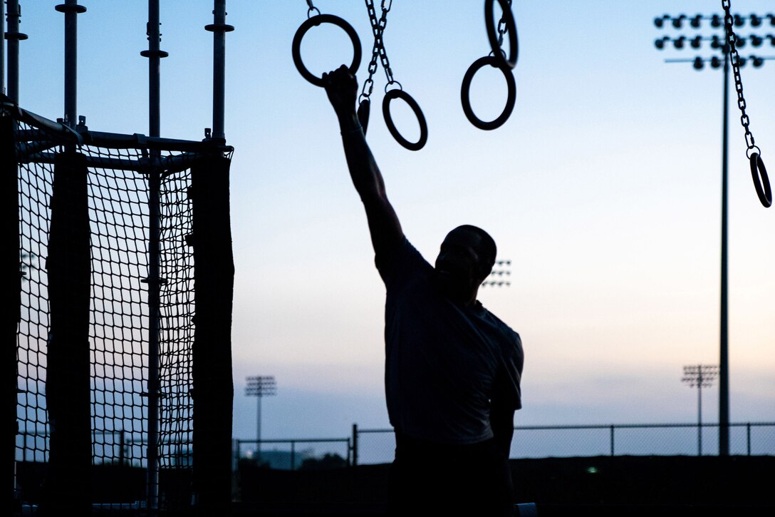 A soldier swings on a circular ring obstacle course with a netted area to the left as shown in silhouette.