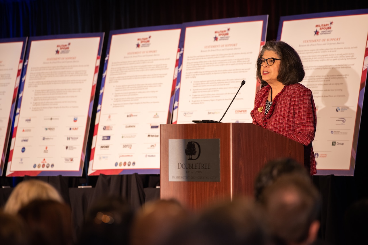 A person speaks at a lectern with several posters in the background.