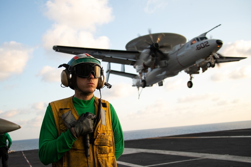 A sailor wearing flight deck equipment stands at the ready as an aircraft lands on the flight deck of an aircraft carrier.