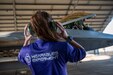 In the foreground a woman stands on a tarmac with he rback to the camera, she has protective  headphones over her ears. She is wearing a shirt that says "Wearables Experiment" and in the background there is a fighter jet under an open airplane hangar.