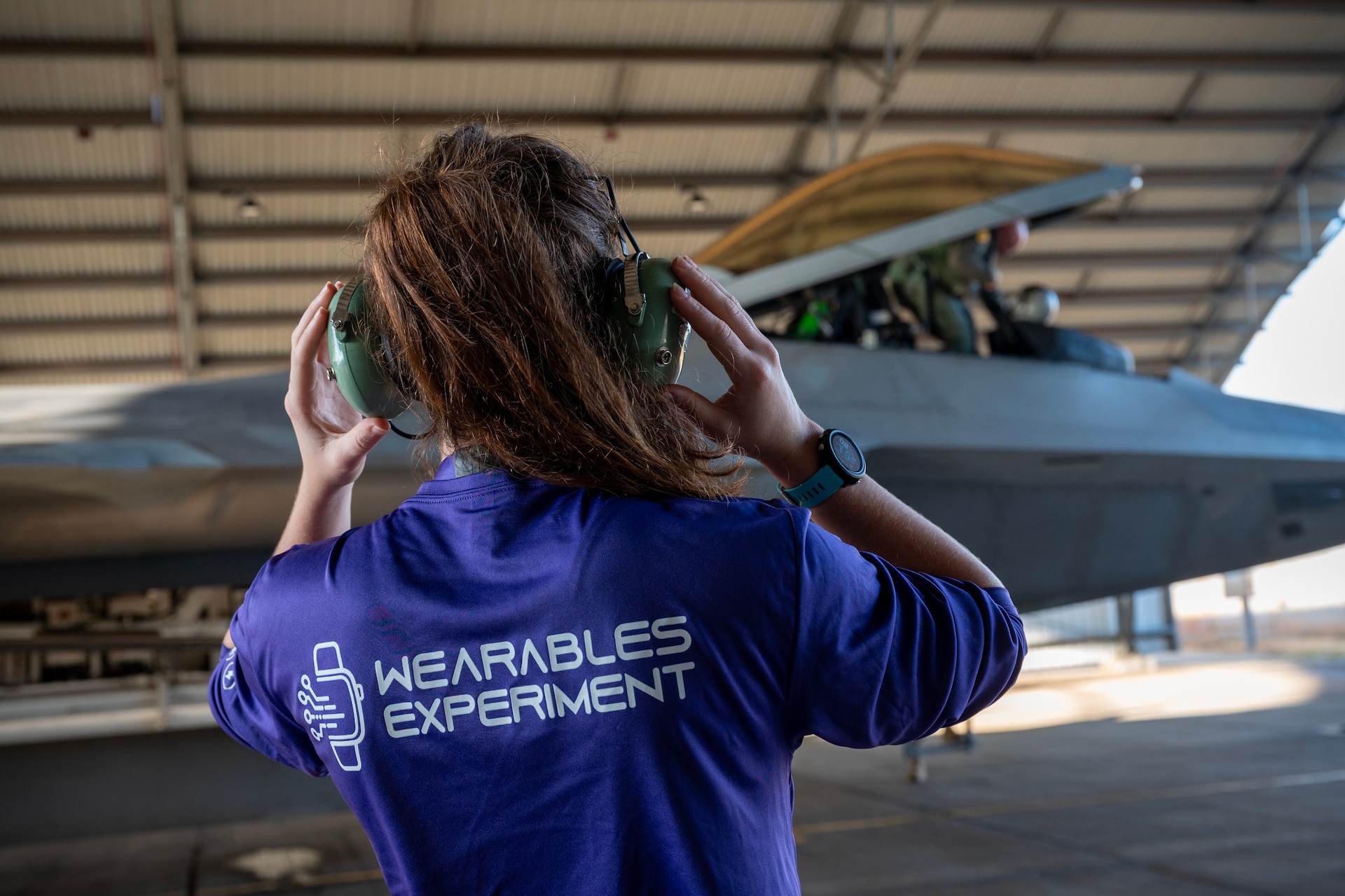 In the foreground a woman stands on a tarmac with he rback to the camera, she has protective  headphones over her ears. She is wearing a shirt that says "Wearables Experiment" and in the background there is a fighter jet under an open airplane hangar.