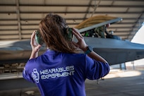 In the foreground a woman stands on a tarmac with he rback to the camera, she has protective  headphones over her ears. She is wearing a shirt that says "Wearables Experiment" and in the background there is a fighter jet under an open airplane hangar.