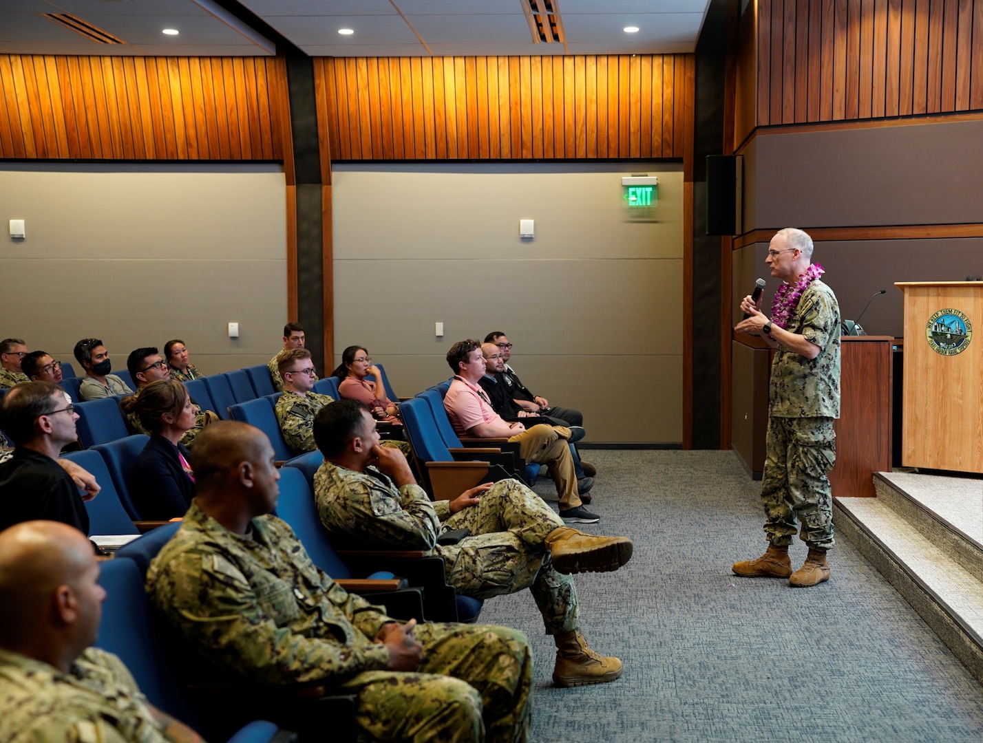 Rear Adm. William Greene, right, stands at the front of an auditorium speaking to a room of Sailors and civilians.